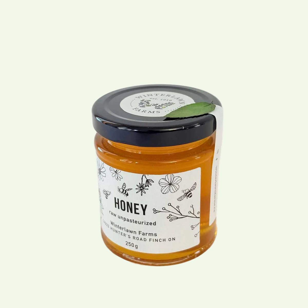 A 250g glass jar of Winterlawn Farms honey, showing the golden amber contents through the clear glass. The jar has a white paper label with black text and illustrations of bees and flowers, and is sealed with a black lid that has a small green leaf accent.