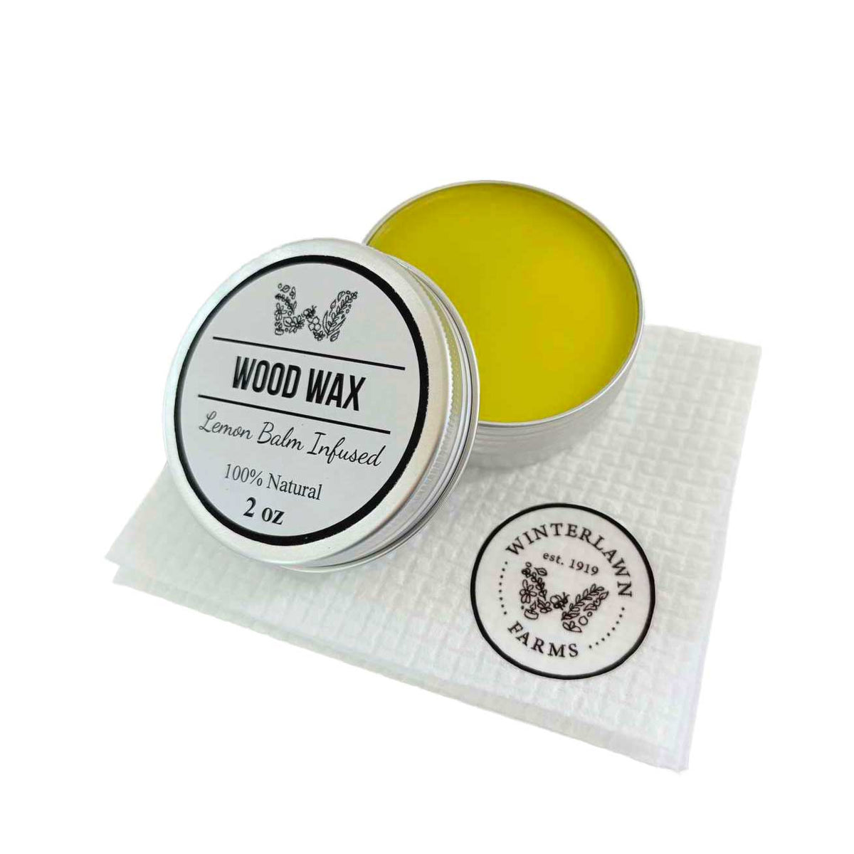 A 2 oz silver tin of Winterlawn Farms Lemon Balm Infused Wood Wax, shown open with the lid beside it on a white textured paper towel. The wood wax inside is a bright yellow-green color.