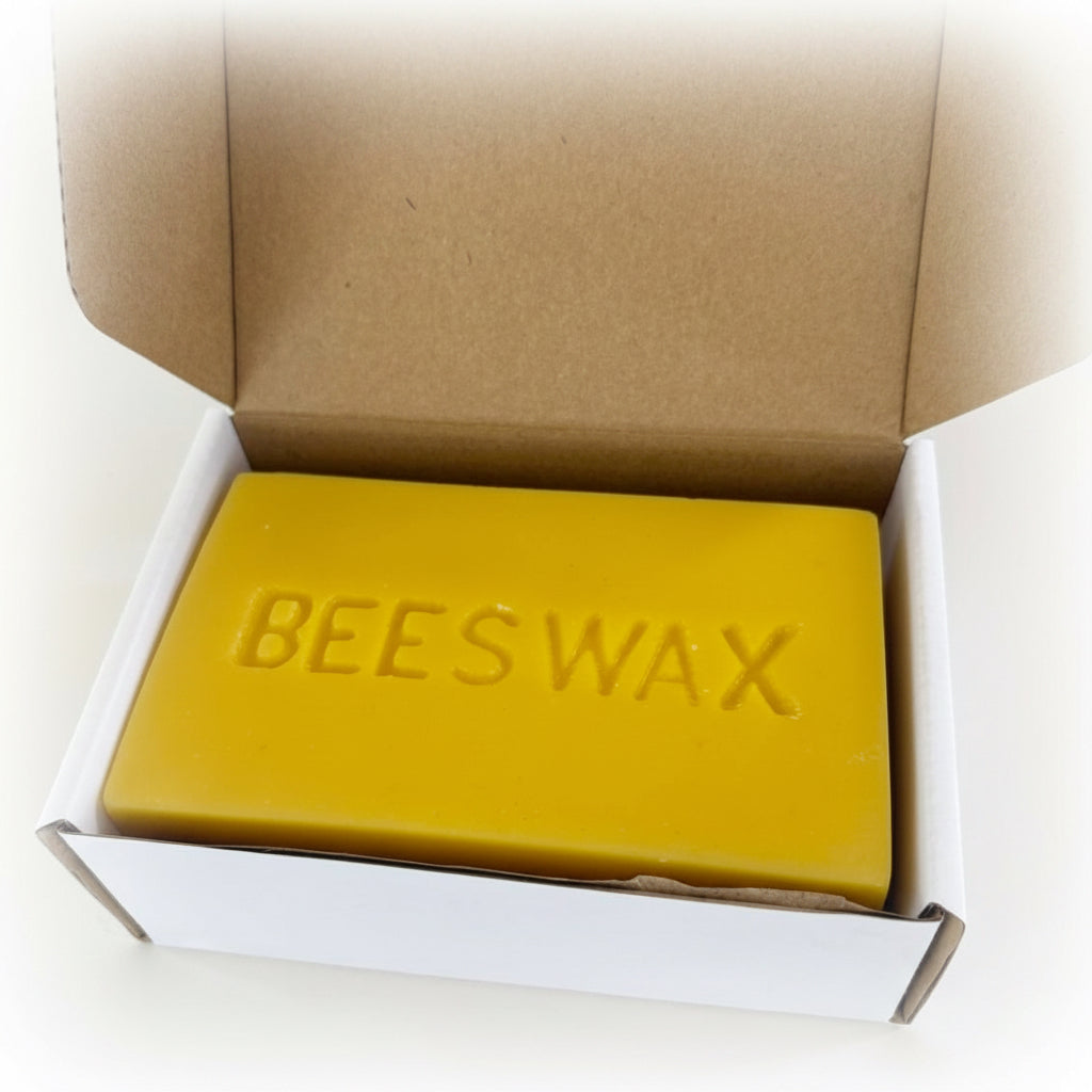 A rectangular bar of yellow beeswax soap embossed with the word 'BEESWAX' on top, presented inside an open brown cardboard box with a white inner base, photographed on a plain light background.