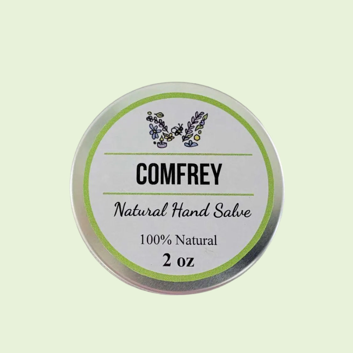 A round silver tin of COMFREY Natural Hand Salve on a plain off-white background. The label is white with a light green ring, featuring the brand name 'COMFREY' and product details '100% Natural 2 oz' with a small botanical illustration.