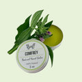 A 2 oz round tin of COMFREY Natural Hand Salve, shown open with the green balm visible, decorated with fresh green leaves and small purple flowers on a clean white background.