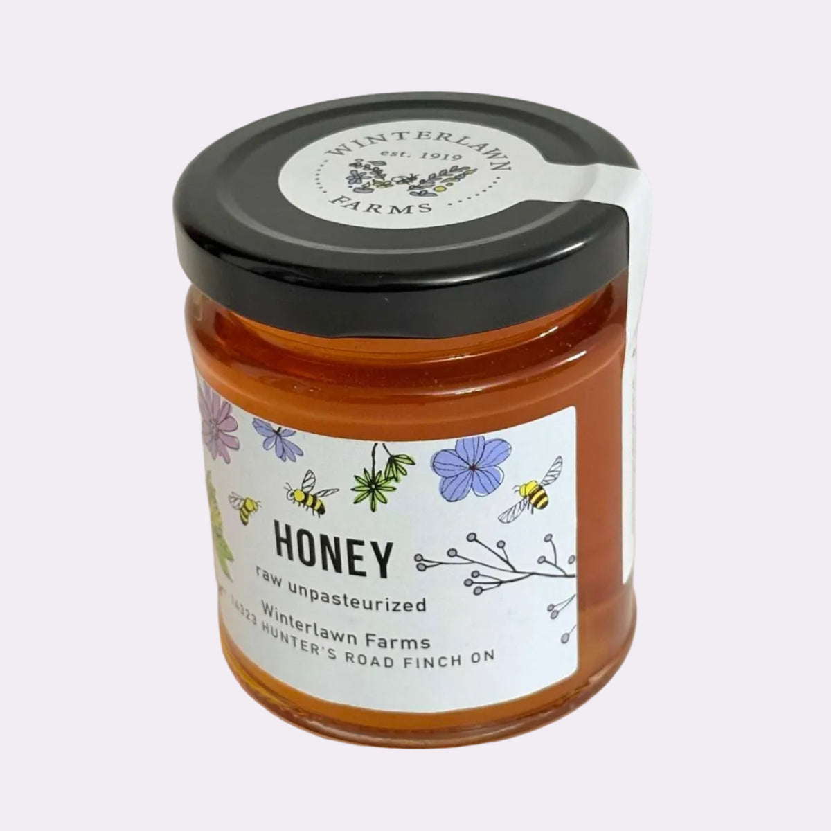 A glass jar of Winterlawn Farms honey with a black lid and a white label featuring floral and bee illustrations, set against a plain light background.