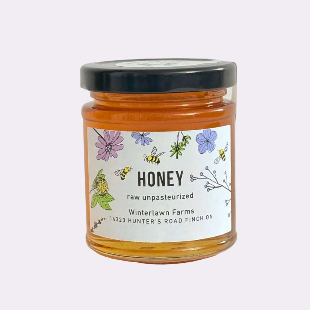 A glass jar of raw unpasteurized honey with a black lid and a white label featuring colorful illustrations of bees and flowers, set on a white surface against a white background.