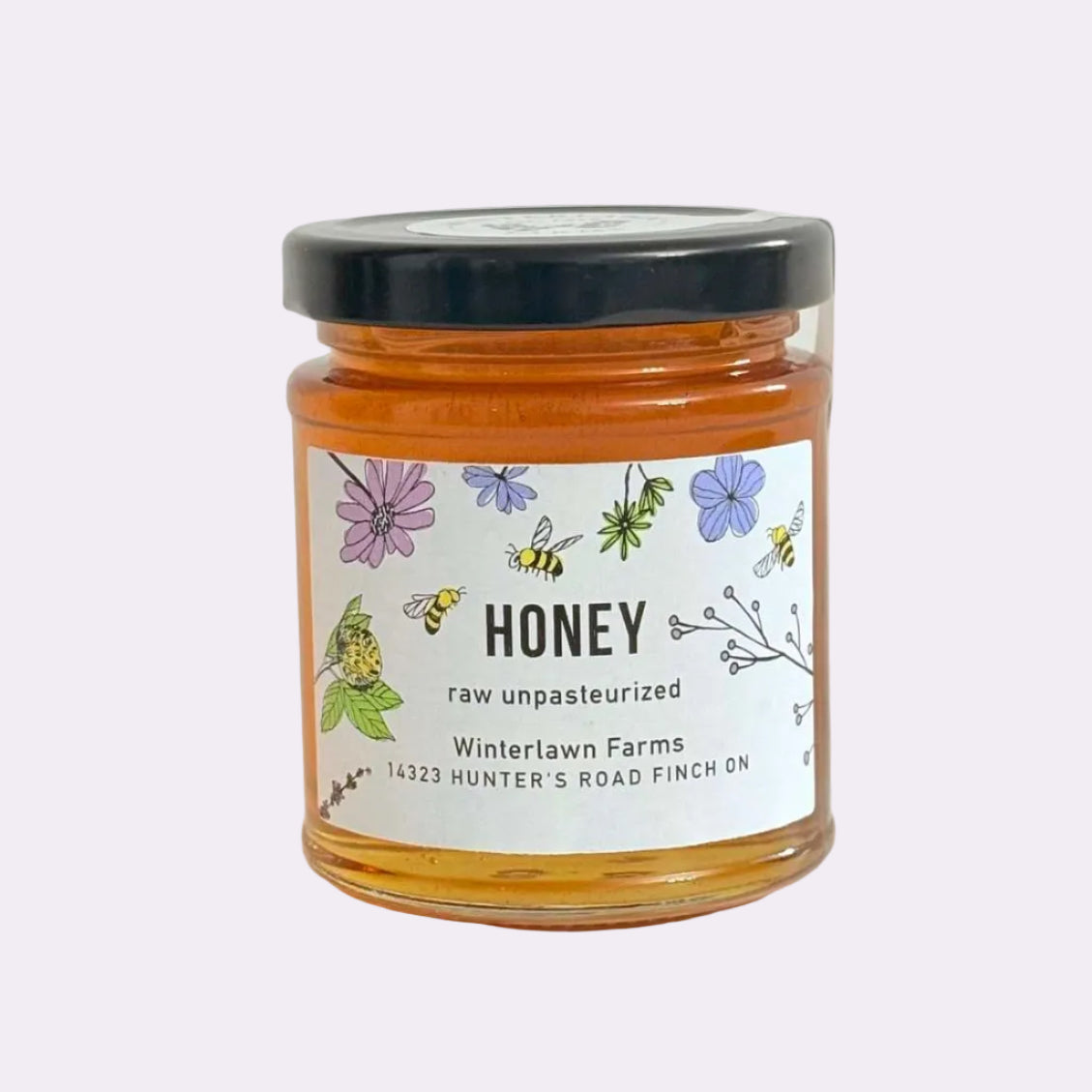 A glass jar of raw unpasteurized honey with a black lid and a white label featuring colorful illustrations of bees and flowers, set on a white surface against a white background.