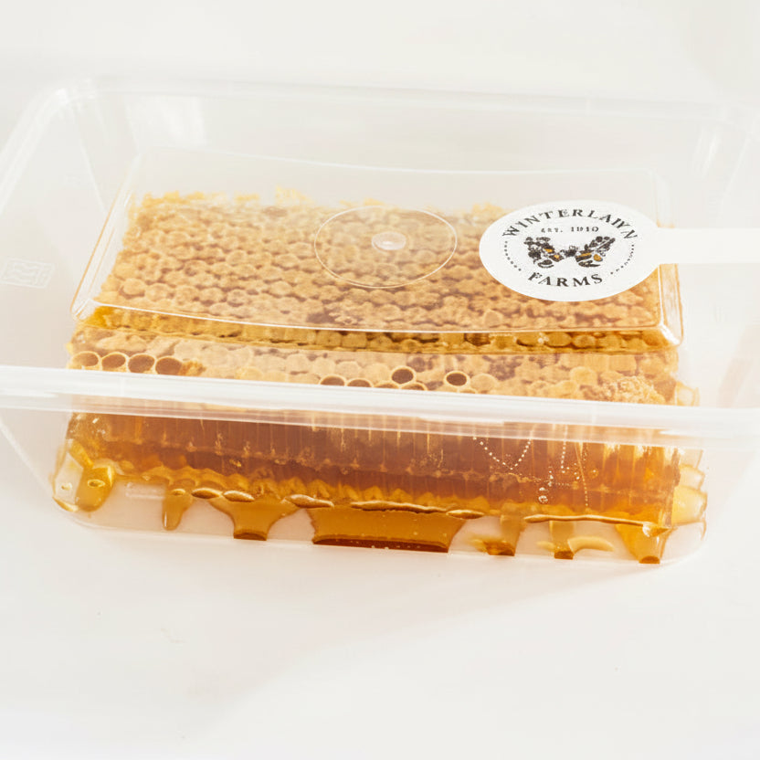 A clear plastic clamshell container holding honeycomb and a strip of raw honey, with a round white sticker label reading 'INTERLAKH FARMS' on a plain light background.