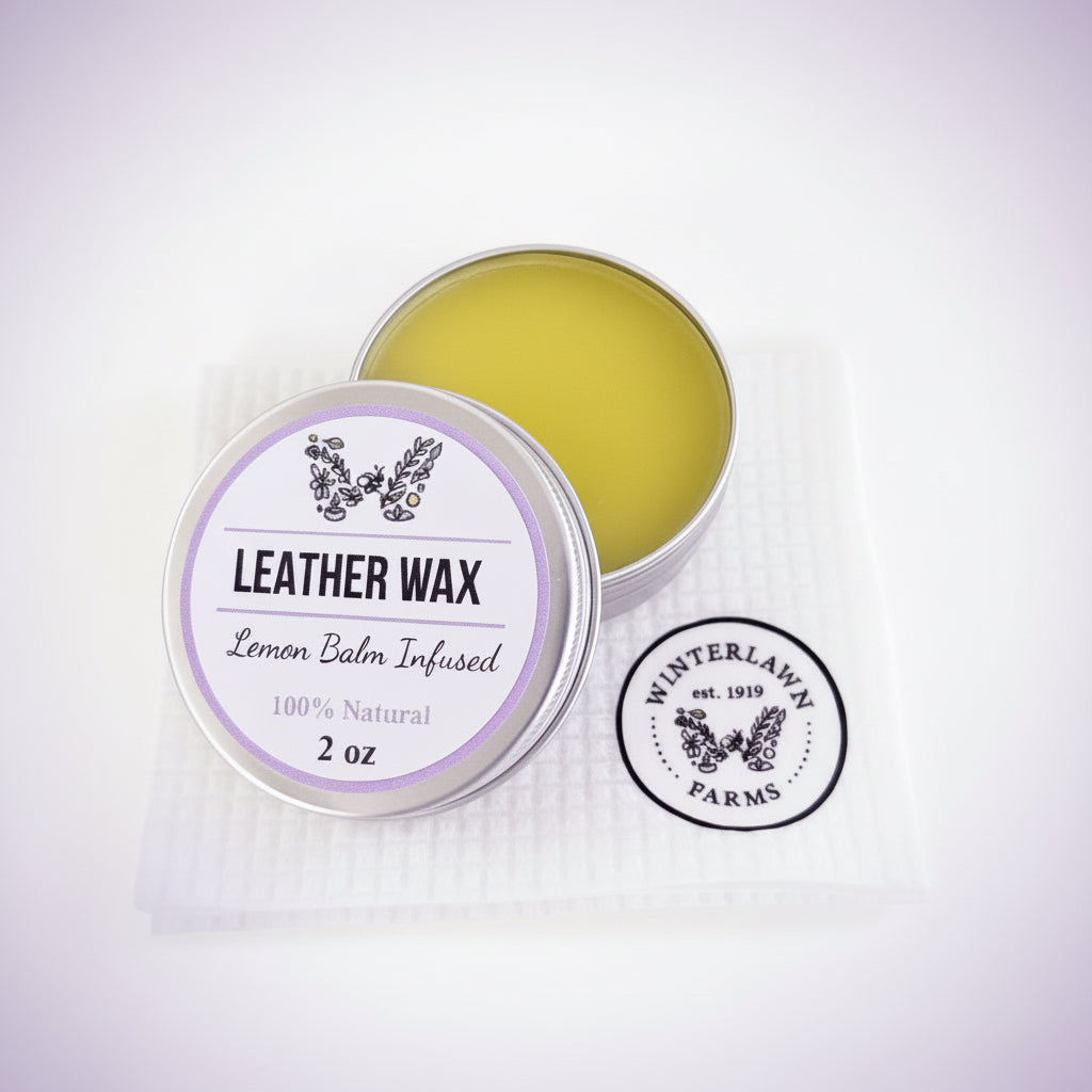 An open round metal tin of leather wax balm sits on a white textured paper towel, with the lid placed beside it. The wax inside is a pale yellow-green, and the lid label reads 'LEATHER WAX' with 'Lemon Balm Infused' and '2 oz.
