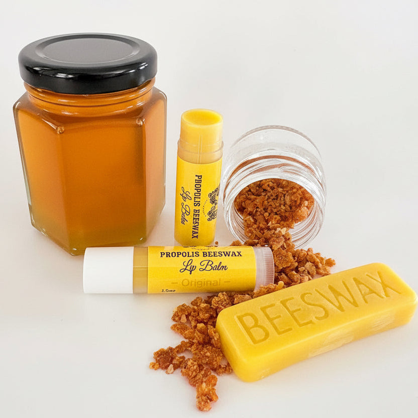 A set of propolis beeswax beauty products on a white background, including a hexagonal jar of golden beeswax with a black lid, a yellow lip balm tube, an open jar of brown propolis crystals, a yellow lip balm bar, and a yellow beeswax bar stamped with 'BEESWAX'.