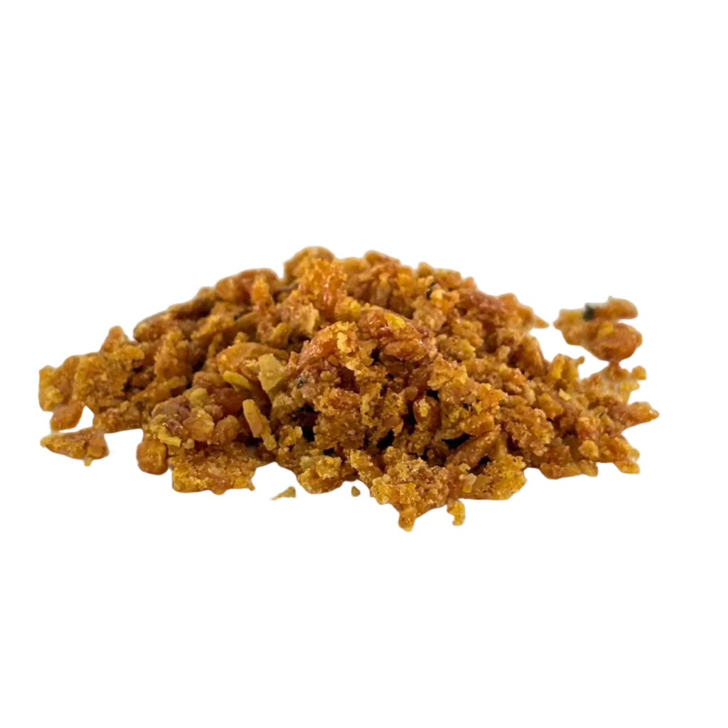 A small pile of golden-brown crumbly snack pieces, possibly crushed nuts or cookie crumbs, placed on a white plate against a light gray background.